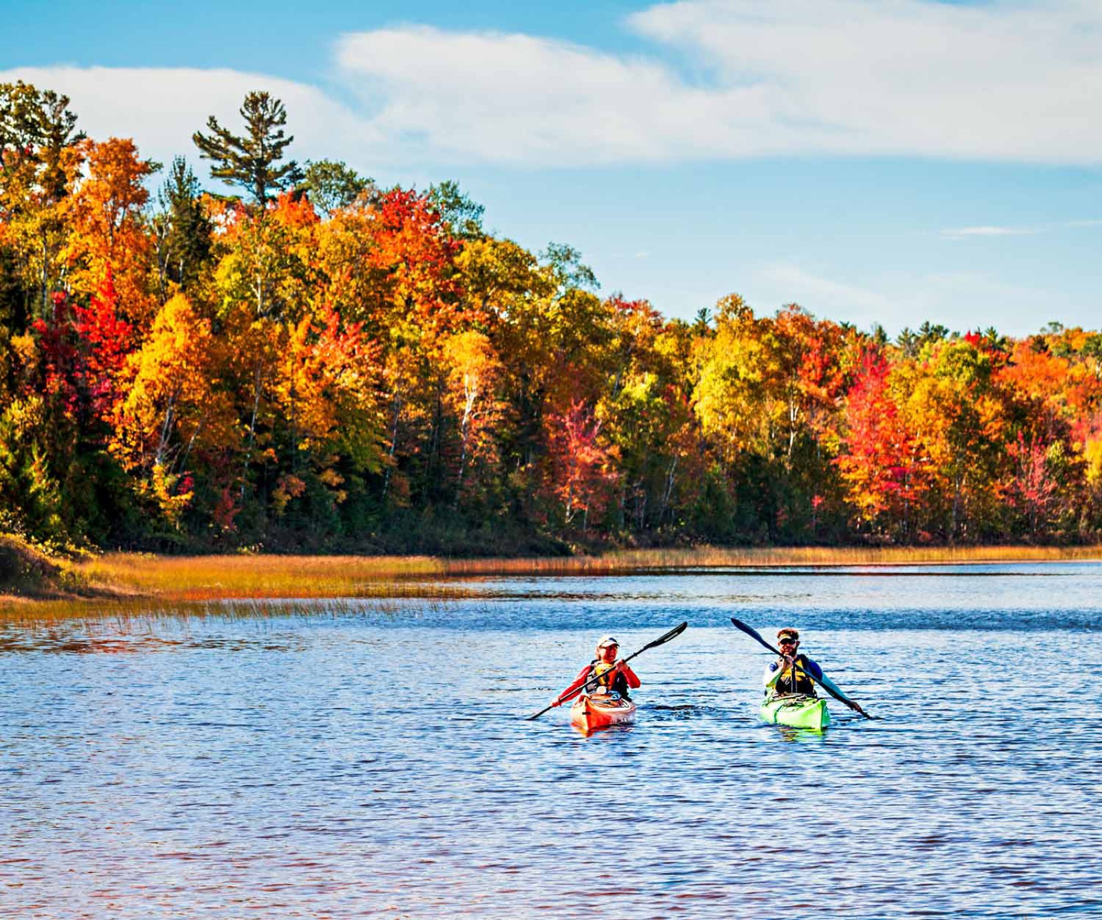 Two kayakers paddling past a shoreline of fall colors